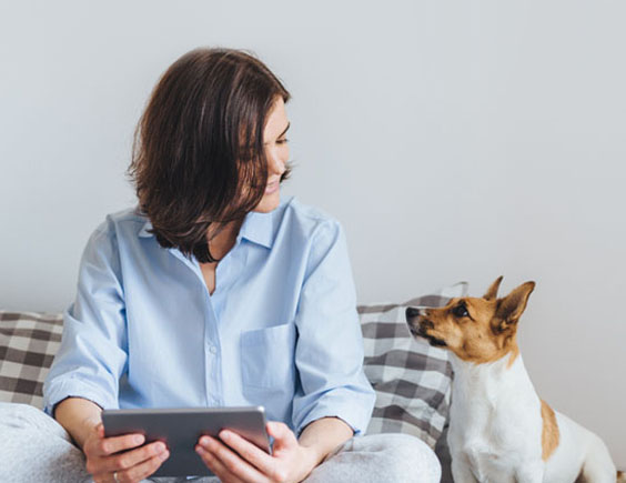 Mujer sentada en una cama sujetando una tablet y mirando a su perro que está al lado