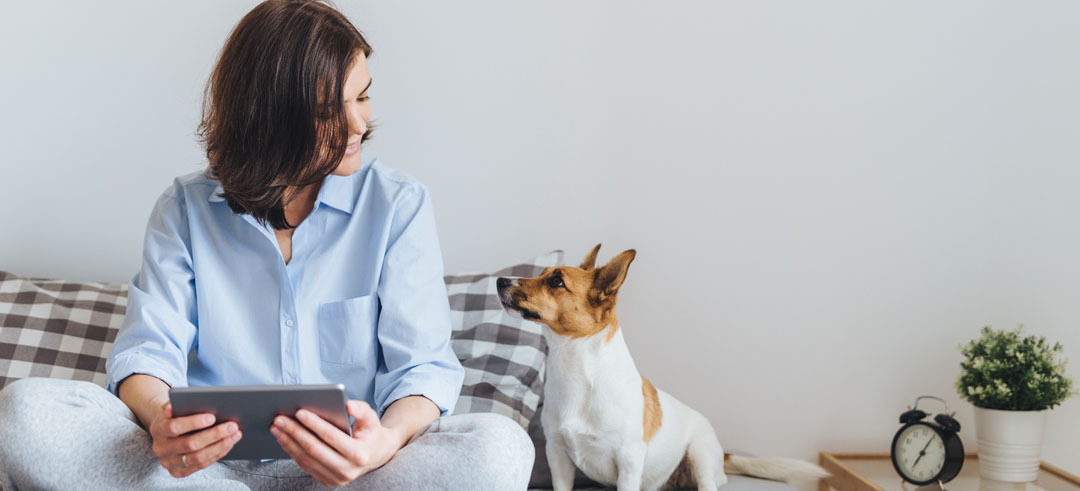 Mujer sentada en una cama sujetando una tablet y mirando a su perro que está al lado